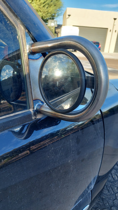 A close-up of a custom welded metal side mirror assembly mounted on the side window of a black vehicle, with an outdoor background visible.